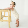 A joyful baby boy in a Makemake Organics Organic Sleeveless Short Romper - Pixie Dots stands holding onto a wooden stool in a bright, white room, looking back with a big smile.