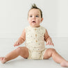 A baby girl with a punk-style hairdo sits on a white surface, wearing a Makemake Organics sleeveless cream-colored Organic Sleeveless Short Romper in Pixie Dots. The baby looks surprised.