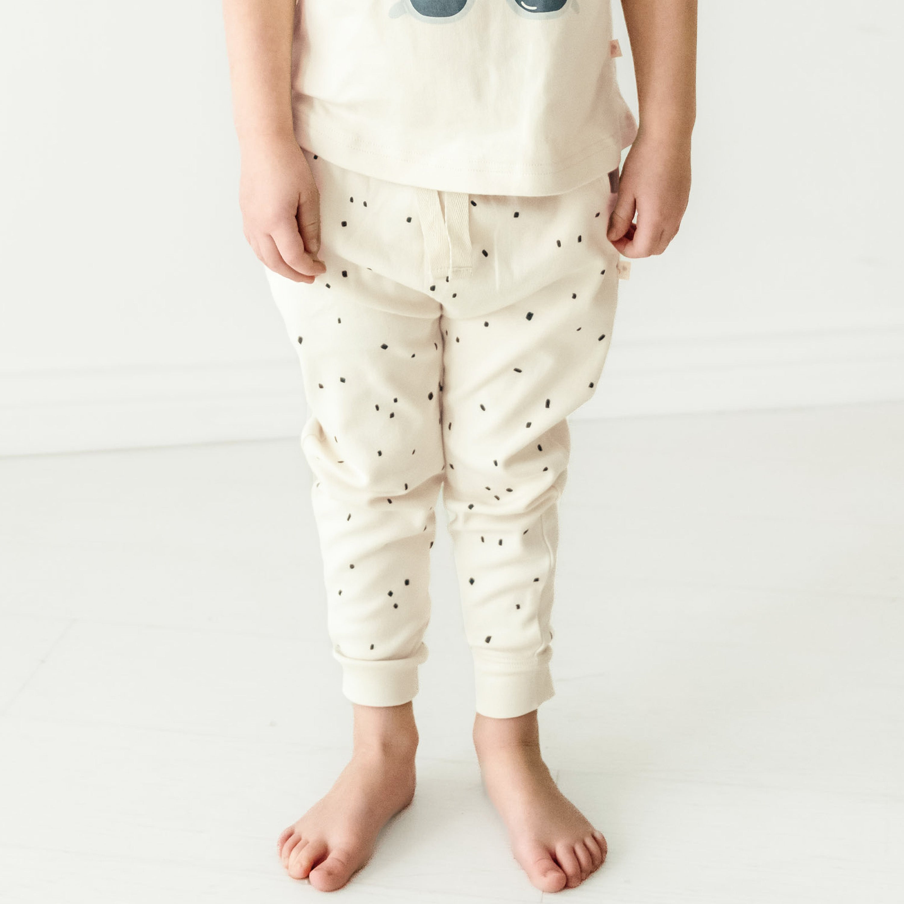 A young toddler standing barefoot, wearing a light-colored shirt with an owl design and Organic Harem Pants in Pixie Dots by Makemake Organics. The background is a clean, white floor and wall.