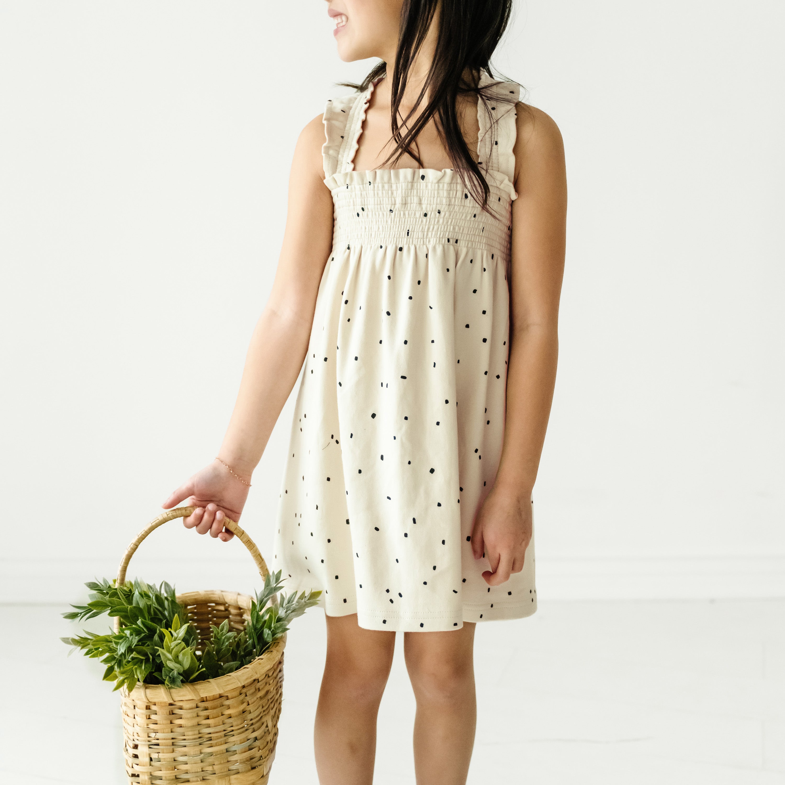 A young girl in a Makemake Organics Organic Smocked Dress in Pixie Dots holds a wicker basket filled with green plants. She is facing away from the camera, showing only a side profile of her toddler-sized frame.
