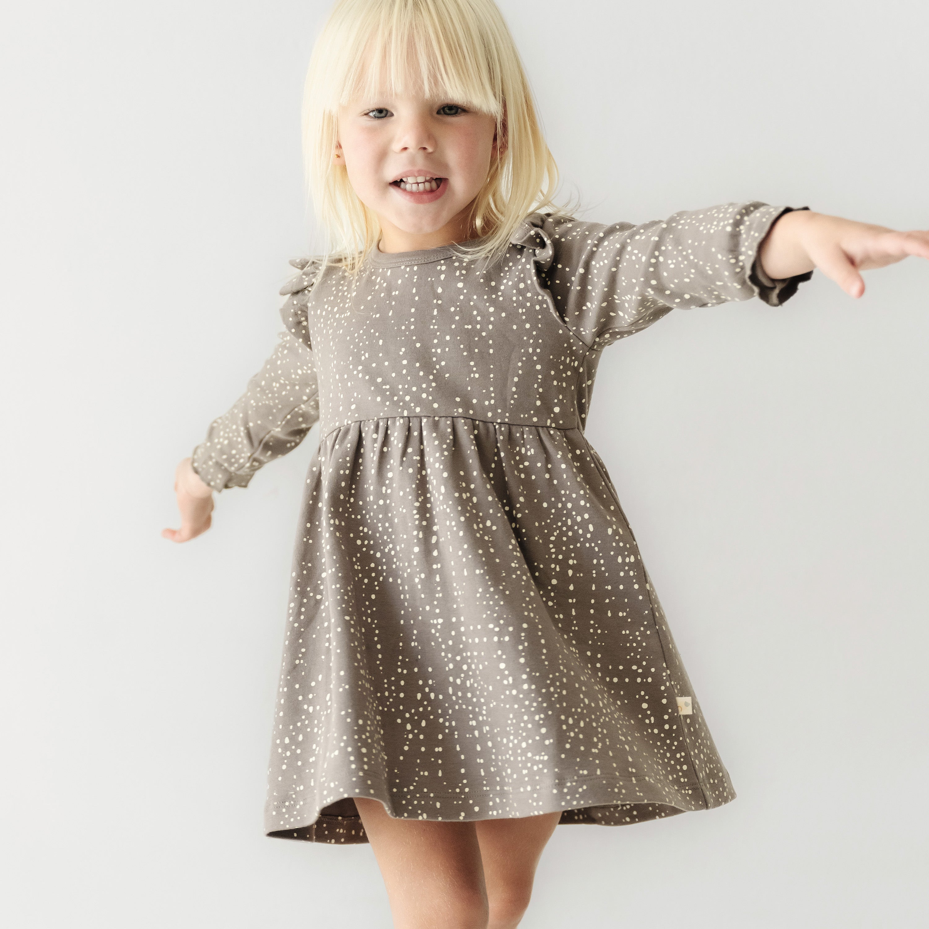 A joyful young girl with blonde hair, wearing an Organic Girls Organic Ruffle Dress - Speckle, playfully reaching out to the side against a light gray background.