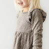 A smiling young girl wearing an Organic Girls Organic Ruffle Dress - Speckle, partially cropped on the left side, stands against a plain white background.