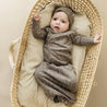 A baby dressed in an Organic Baby speckle kimono knotted sleep gown and hood lies in a wicker bassinet, looking up with a surprised expression.
