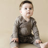 A baby with expressive eyes and dark hair sits on a beige background, wearing a gray Organic Kimono Onesie & Pants Set - Speckle from Organic Baby, looking upward with a curious expression.