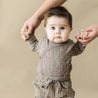 Sentence with replaced product and brand name: A baby with wide eyes and dark hair, wearing an Organic Baby speckled kimono onesie and pants set, is being held up by the hands of an adult against a beige background.