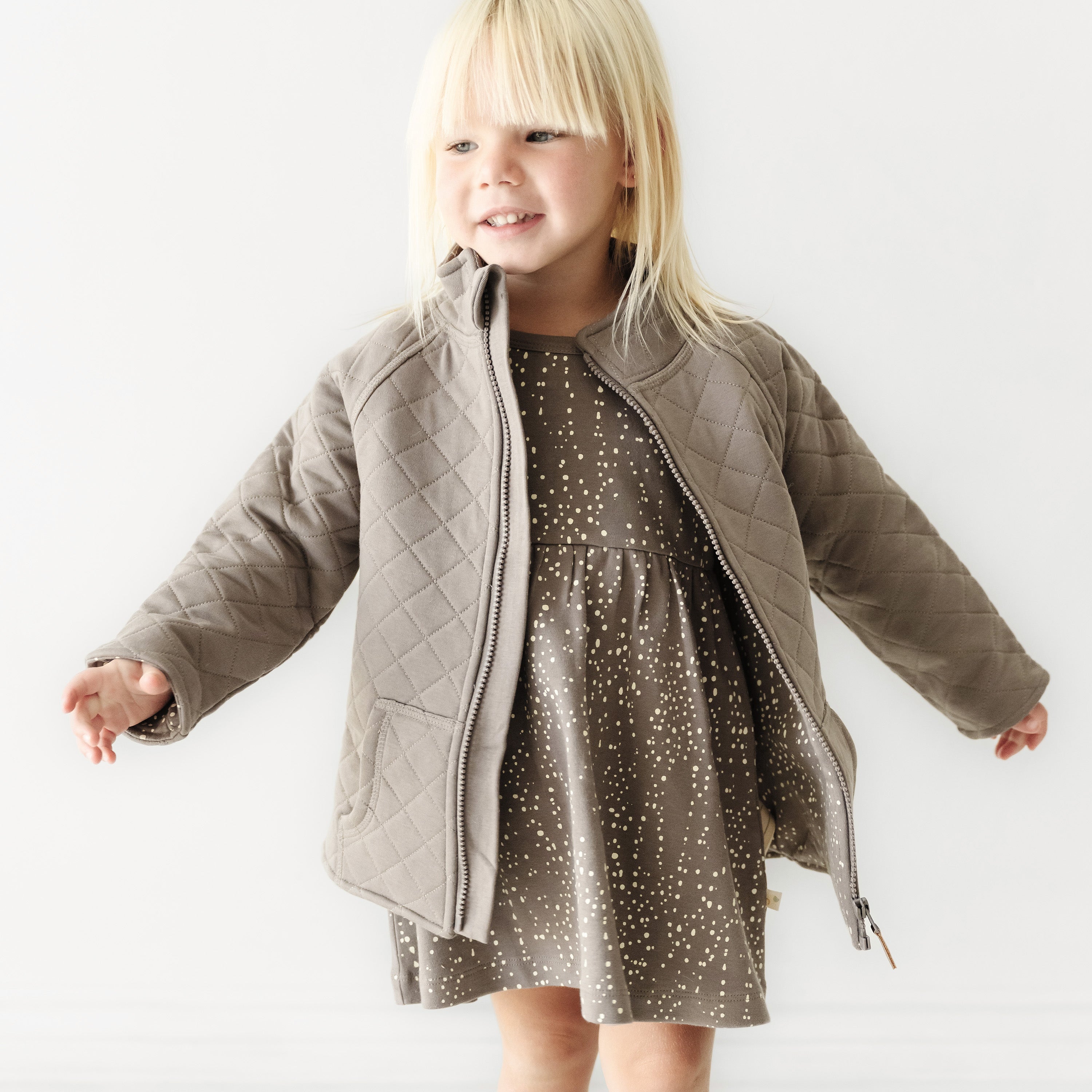 A young child with blond hair smiling and wearing a gray quilted jacket from Organic Kids over a dotted dress, standing against a white background.