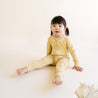 A toddler with short black hair, wearing an Organic Baby Organic 2-Way Zip Romper - Yellowstone with a polka dot pattern, sits on a pale background, looking at the camera with a neutral expression.