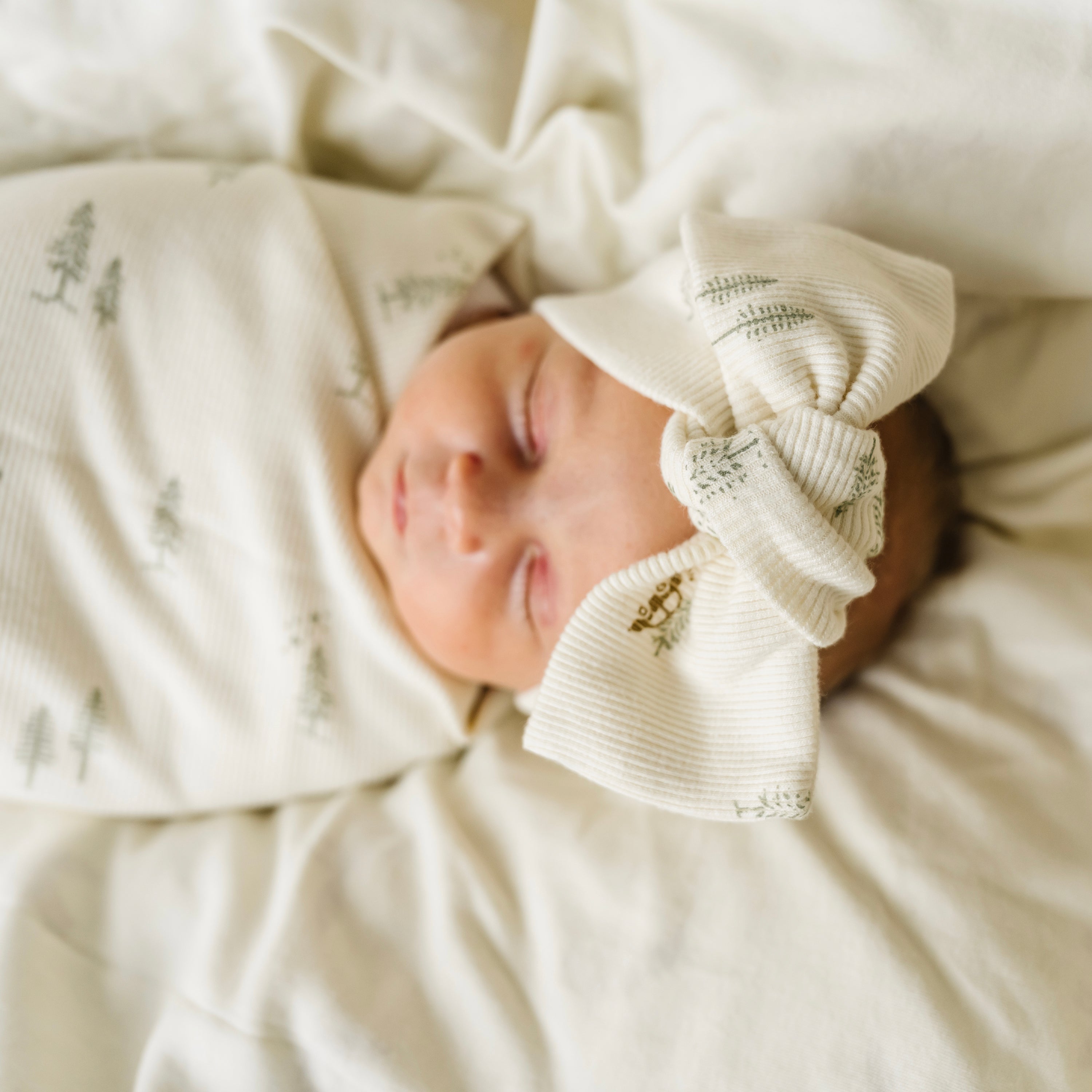 A newborn baby sleeps peacefully on a soft, light-colored Makemake Organics blanket, wearing a white onesie and a cute, knotted hat with leaf patterns. The baby is also bundled up in an Organic Headwrap from Wildwood & Evergreen Fir.