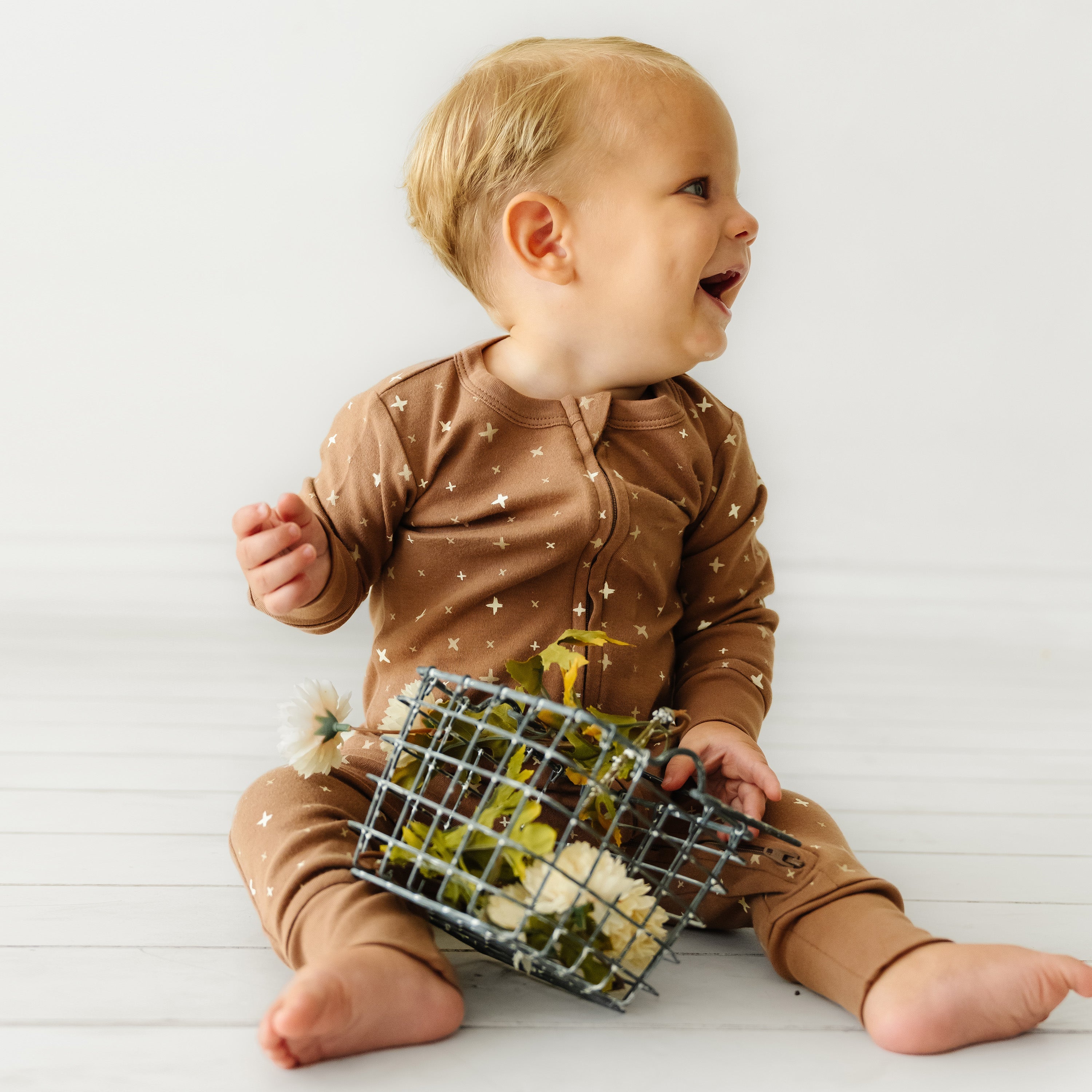 A joyful baby in an Organic Baby Sparkle brown onesie decorated with stars sits on a white background, holding a small wire basket filled with yellow flowers, looking off to the side with a bright smile.