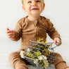 A joyful toddler in an Organic Baby brown star-patterned onesie sits on a white floor, playfully holding a small wire basket filled with yellow flowers.